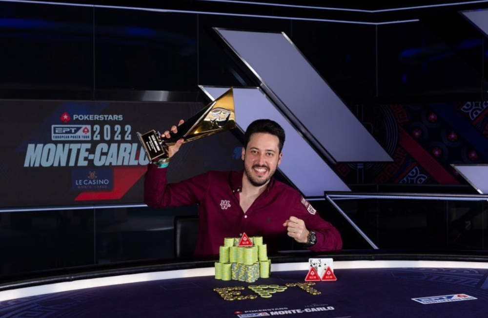 Adrian Mateo Diaz is seen sitting behind a desk holding up his EPT Monte Carlo Super High Roller trophy and smiling. A pile of chips are in front of him.