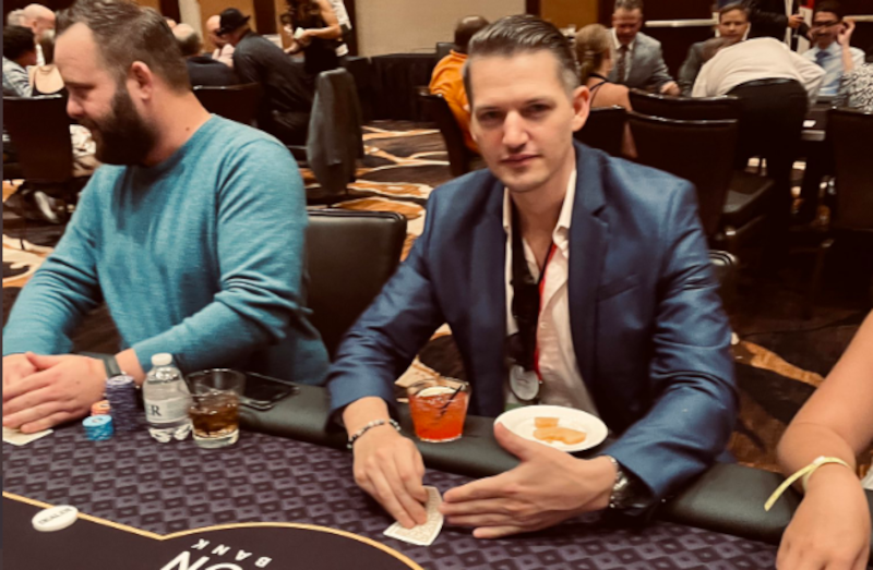 Joey Ingram is seen sitting at a poker table. He's wearing a slick dark blue shiny blazer with a button-down shirt under it. his hair is slicked back. in front of him is a drink and a plate with a snack.