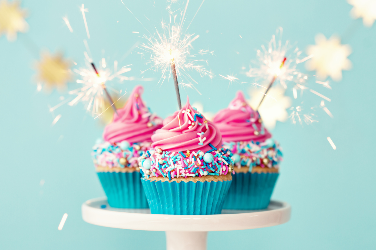 3 cupcakes with sparklers and pink frosting on a cake stand in front of a blue background.