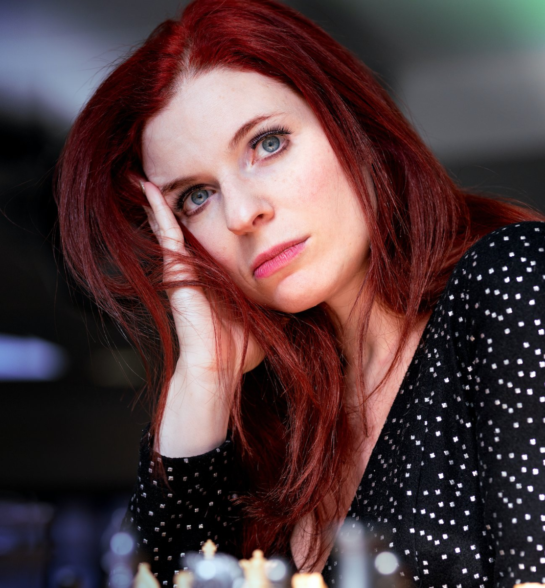 Author headshot of Poker & Chess phenom Jennifer Shahade, who signed two-book deal to write about being a top female chess player . She has straight dark red hair, serious blue eyes, and is wearing a black top with a pattern of small white squares. 