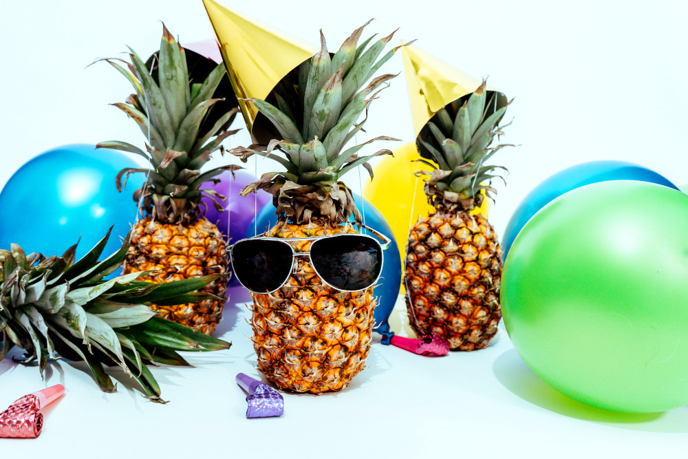 Three pineapples are seen partying. The one in the middle wears aviator sunglasses. All 3 wear party hats. Around them are balloons and kazoos. BetMGM, bwin, and partypoker bring the party to the Ontario online poker scene.