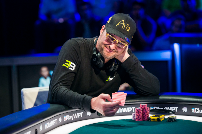 Phil Hellmuth -- a white male wearing square glasses, a black long sleeve shirt, and a black ARIA dad hat -- sits at a WPT branded poker table, smiling as he looks down at his cards, a pile of chips in front of him.